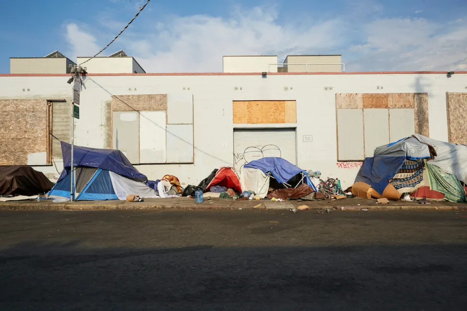 Small tents set up on sidewalk by unhoused people next to boarded-up one-story industrial building in San Francisco, California.